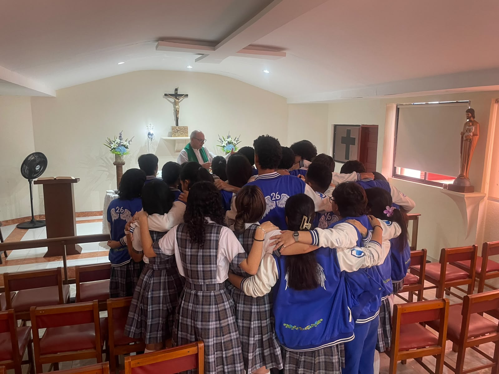 Estudiantes de la promoción 2026 abrazados en círculo dentro de la capilla, acompañados por el sacerdote junto al altar